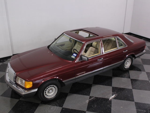 Front quarter view of a Cabernet Red 1985 Mercedes-Benz 500SEL with US-spec headlights and chrome wheels, parked outdoors.