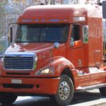 Orange Freightliner Columbia semi-truck transporting logs, featuring headlights also used in Mercedes-Benz ML-Class vehicles.