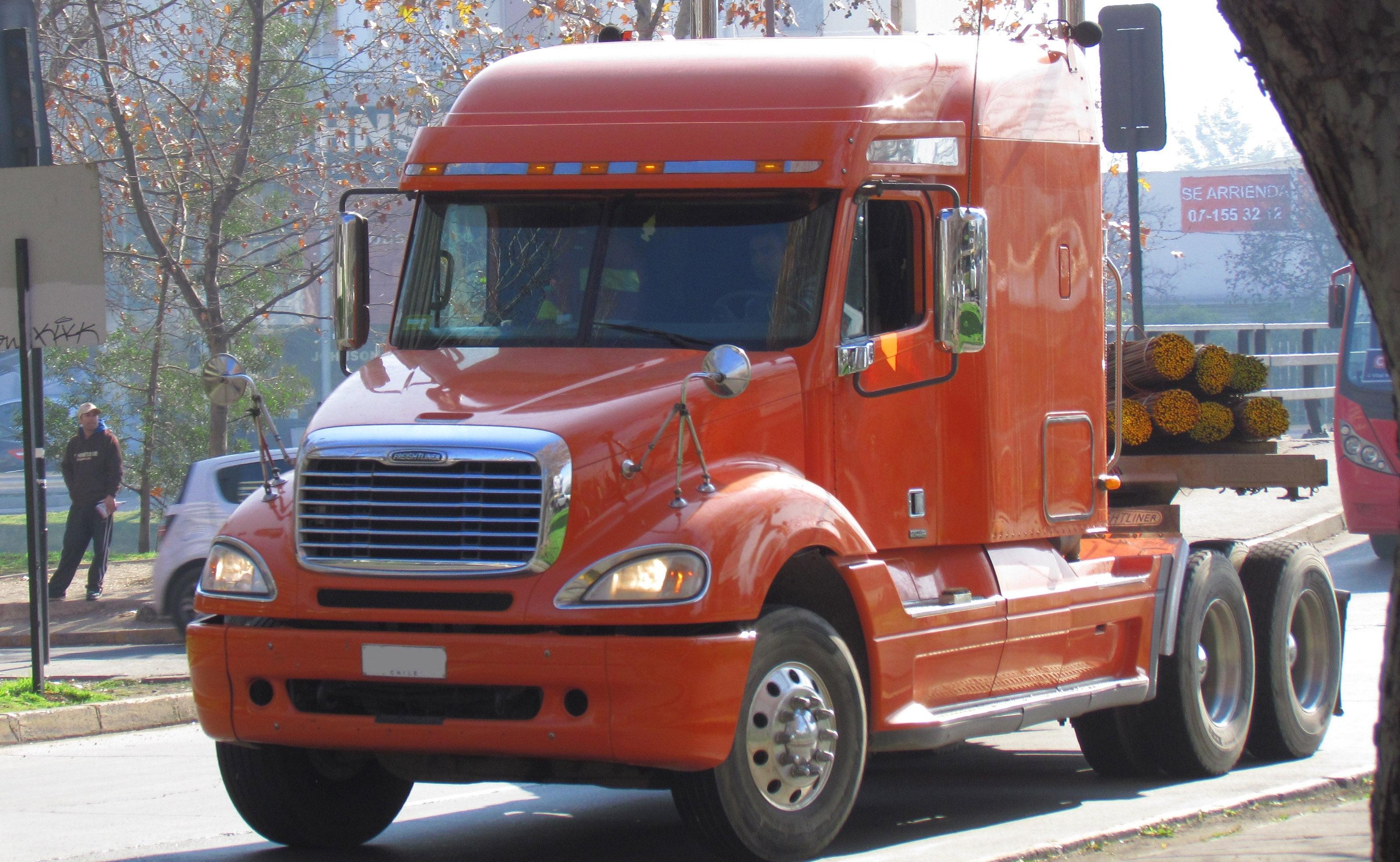 Orange Freightliner Columbia semi-truck transporting logs, featuring headlights also used in Mercedes-Benz ML-Class vehicles.