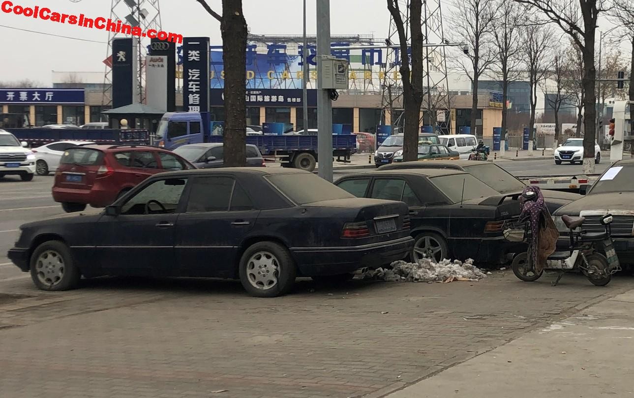 Collection of black Mercedes-Benz cars parked on a snowy pavement in front of car repair shops, suggesting an abandoned Mercedes dealership.