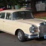 Front three quarter view of a black 1965 Mercedes 220S W111 with wire wheels parked on a street.