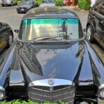 Black Mercedes-Benz Fintail W110 driving on a road, partially obscured by a hedge, hinting at its elusive nature