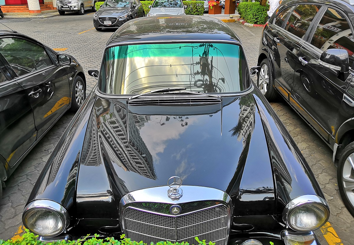 Black Mercedes-Benz Fintail W110 driving on a road, partially obscured by a hedge, hinting at its elusive nature