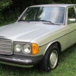 Front side view of a faded red 1980 Mercedes-Benz 300D with fog lights on a paved lot, showcasing its weathered paint condition.