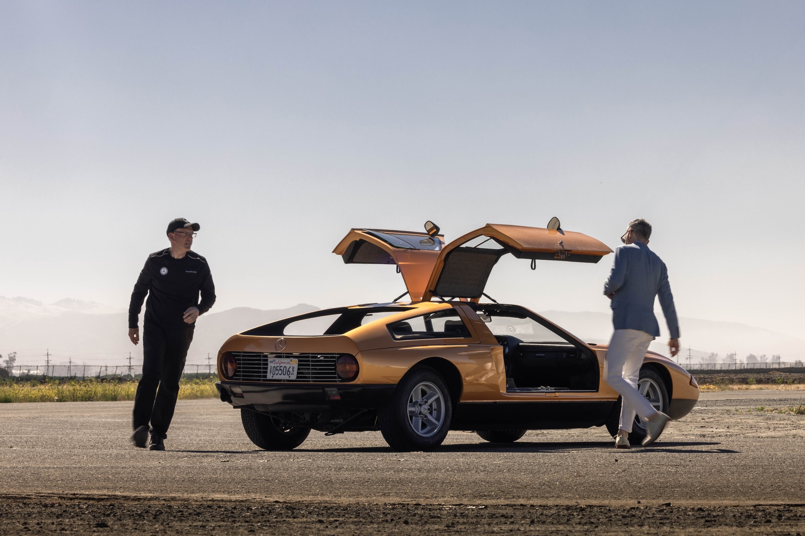 Mercedes-Benz C111 concept car in orange, parked on an airport tarmac with private jets in the background, showcasing its wedge shape and classic design.